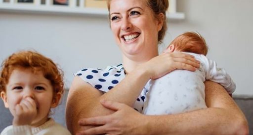 A joyful woman with auburn hair in a polka-dot top cradles a newborn baby in a white sleeper. To her left, a toddler with red, curly hair smiles with a hand near their mouth, wearing a patterned cream sweater. They are indoors with framed pictures on the wall behind them.